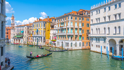 Brightly colored buildings line the Canal Grande as gondolas glide through the water under a clear blue sky. Tourists enjoy the vibrant atmosphere of Venice during a perfect day.