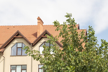 Close-up photograph of the roof and facade of a beige-colored, modern house with a red tile roof, surrounded by trees in the foreground