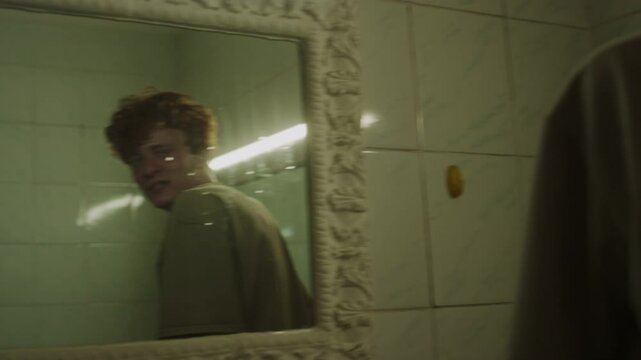 Young man turning between two mirrors in public bathroom looking in one with mad smile and in other with fear, then leaning on wall corner and sliding down