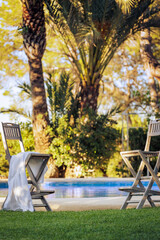 Meeting place by the pool.. Two high bar stools with a carelessly left towel, by the pool on the green lawn against a backdrop of palm trees lit by the setting sun.