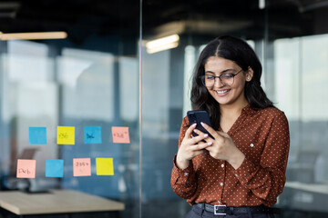 Young business woman using phone inside office, company worker smiling typing text message and...