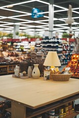 An empty wooden table top with a blurred supermarket background.   There are various items like a vase, a basket and a lamp on the table. 