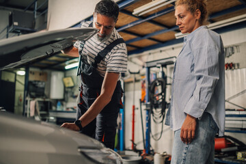 Fototapeta premium Customer and technician looking under car hood at mechanic shop.