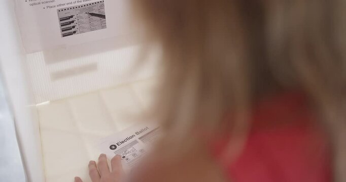 Close, over the shoulder view of young woman voter filling in a ballot at a voting booth in the USA. Hand held camera on stabilized gimbal. Candidate names obscured. 4K DCI