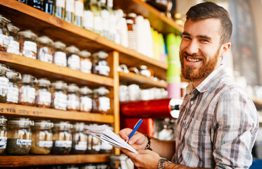 Man, writing and shop owner in portrait at cafe by shelf, paperwork and inspection for stock with smile. Person, entrepreneur and documents with notes, inventory and restaurant management in Turkey