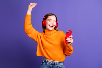 Photo of blonde cheerful teenager girl in orange turtleneck and jeans dancing with smartphone...