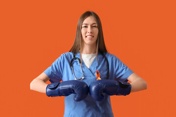 Beautiful young happy female doctor in boxing gloves with ribbon on orange background. Multiple...