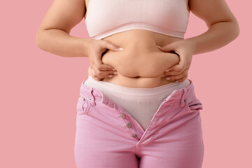 Overweight young woman in tight jeans touching her fat belly on pink background, closeup. Weight...