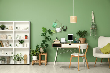 Stylish workplace with tablet on stand, shelving unit and different houseplants near green wall at home