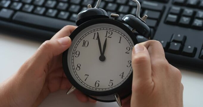 Businesswoman in an office sitting behind a computer and holding an alarm clock in her hands that is showing few minutes until the deadline. Close up.