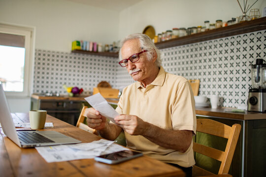 Elderly man managing bills and finances on laptop in kitchen