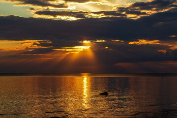 A serene sunset casting golden hues over calm sea waters, with clouds parting to reveal the sun’s...