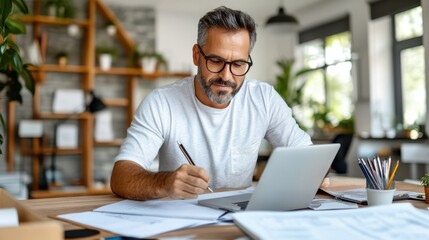 A man wearing glasses, focusing deeply on his work, writes on paper while using a laptop in a home office surrounded by plants and natural light.