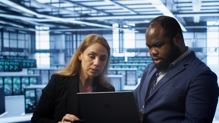 Teamworking colleagues in server farm using laptop to patch network issues affecting transfer rates. Coworkers in data center doing brainstorming, fixing errors affecting supercomputers, camera A