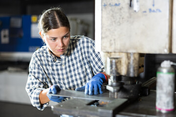 Focused young female worker operating hydraulic punch press in manufacturing facility, creating precise holes in metal sheets..