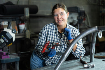 Young girl, professional technician with clamp in hands, wearing checkered shirt and gloves, posing in industrial metal workshop, smiling confidently and friendly at camera..