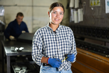 Young female worker in uniform posing with tape measure in metallurgical workshop