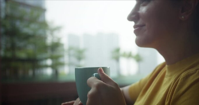 Attractive peaceful dreamy woman with curly hair wearing glasses holding cup enjoy favorite tea drink morning invigorating coffee beverage while, the sun shines on her from the big window.