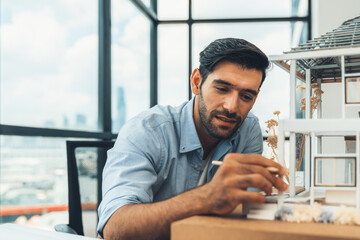 Closeup portrait of smart caucasian architect engineer inspect house model while thinking about building construction at architect studio on table with project plan, blueprint, equipment. Tracery.