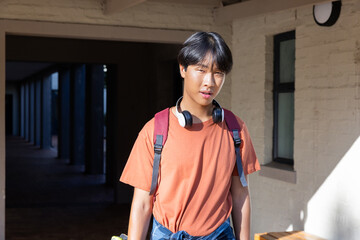 Obraz premium Asian teenage boy with headphones and backpack near school building, looking ahead