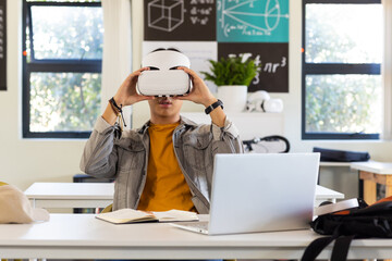 In school, asian teenage boy using VR headset while studying with laptop and notebook