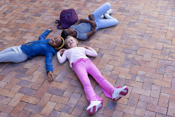 Lying on brick pavement, three multiracial girls relaxing with backpack nearby at school