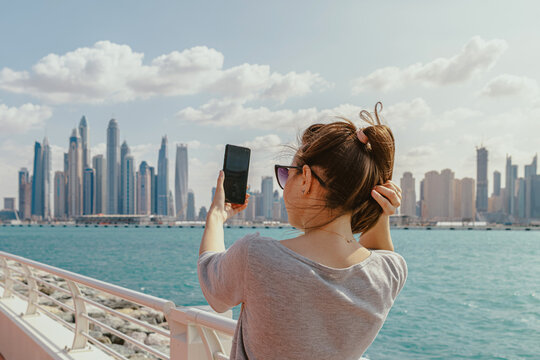 Young woman taking selfie by the waterfront on her smartphone with a city skyline on the background