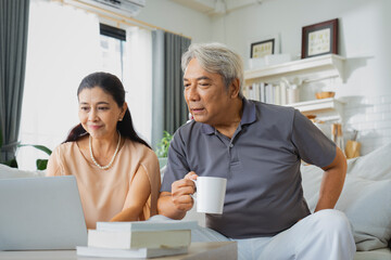 Senior Asian man and woman sitting on sofa looking at travel details on laptop inviting each other...