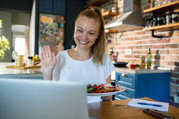 Smiling woman eating salad while video chatting from home kitchen