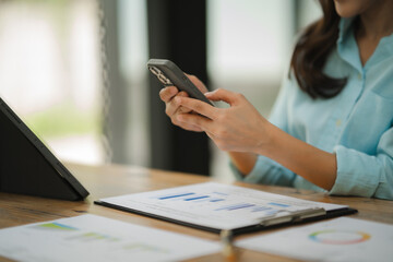 Businesswoman Checking Phone: A close-up image of a businesswoman's hands holding a smartphone, reviewing financial data on a tablet and clipboard, showcasing the seamless integration of technology in