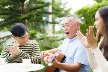 Happy Asian family spending time together, man and woman let their father playing a guitar and singing in backyard garden at home. Mental health care and wellbeing in senior adult.