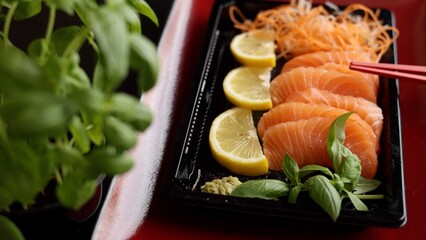 Sashimi displayed on elegant red plate captures essence of refined dining. Lemon, basil, and soy sauce enhance vibrant flavors, while red sticks provide finishing touch for perfect presentation.