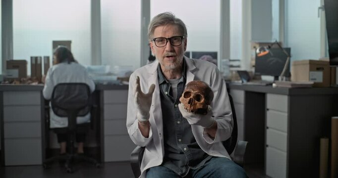 Mature lab worker or scientist speaks to camera, shows fossil skull of ancient human during interview for documentary film in archaeological laboratory. Colleague works in the background. Static Shot.
