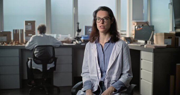 Female young scientist in lab coat talks to the camera and gives interview for popular science film while sitting in archaeological scientific lab. Male colleague works in the background. Static Shot.
