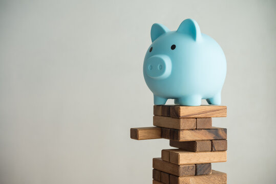 Blue piggy bank on unstable wood tower block game with white wall background copy space. Risk and personal financial management, money savings in economy crisis, retirement planning, banking concept.