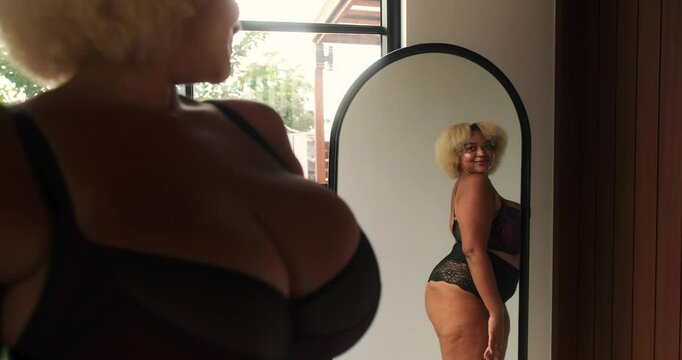 Young African American plus size curvy woman stands in front of the mirror in her underwear and admires her reflection. Self love and acceptance, body positive. Seductive female body.