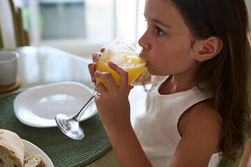 Young girl enjoying a refreshing glass of orange juice at breakfast table on a sunny morning