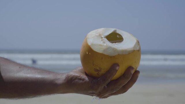 A fresh king coconut is expertly cut open on a sunlit beach, revealing its hydrating water amidst the sandy shoreline. The scene captures the vibrant, natural process against a stunning coastal beach.
