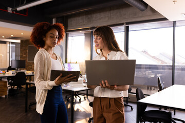 Discussing project, two multiracial women holding laptop and tablet in modern office