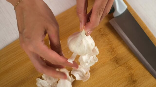 Woman cooking peeling garlic using knife on wooden board, hands close-up. Preparing food, meal on kitchen at home. Cooking dish. Culinary, cuisine, cook with fresh products, ingredients concept.