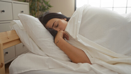 Young woman sleeping in a bed, wrapped in white sheets, in a cozy bedroom at home with a dresser in the background.