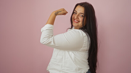 Young curvy hispanic woman flexing muscles isolated over pink background looking confident and...