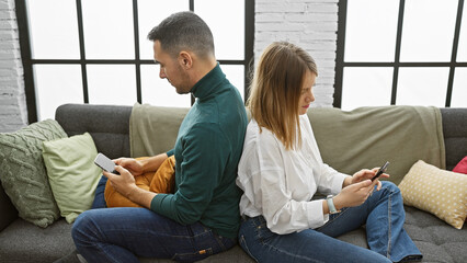 A man and woman sit back-to-back on a sofa, absorbed in their smartphones, in a cozy, well-lit living room.