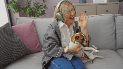 Middle-aged blonde woman in a living room interacting with a small dog on a sofa while wearing headphones and waving hello indoors.