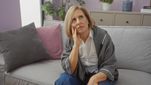 Mature blonde woman sitting pensively on a couch in a cozy living room, adorned with cushions and plants in the background.