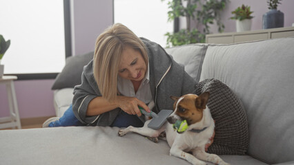 A mature blonde caucasian woman grooms her dog on a cozy living room sofa with plants in the backgrounds