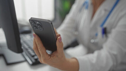 A mature female doctor in a hospital room holds a smartphone in one hand while working at her computer with the other, epitomizing a modern healthcare professional's workplace.