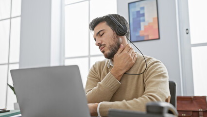 A young hispanic man with a beard grimacing in pain as he touches his neck in an office setting, signifying discomfort while using headphones and working on a laptop.