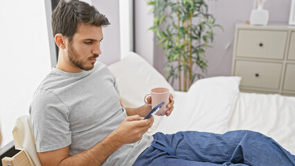 A young hispanic man holding his phone and a mug while sitting in bed within a cozy bedroom interior.
