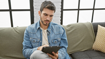 Handsome hispanic man with beard focuses intently on tablet in modern living room setting